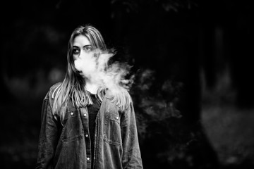 Vape teenager. Young white addict caucasian teenage girl in casual clothes vaping electronic cigarette with cannabis liquid near the tree in the forest in the autumn evening.  Black and white.