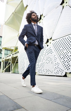 Successful black employee in trendy suit walking along street with hands in pockets and looking away