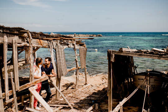 Enamored man and woman hugging on shore under shabby wooden canopy in summer and looking at each other