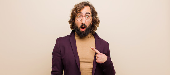 young bearded crazy man looking shocked and surprised with mouth wide open, pointing to self against flat color wall