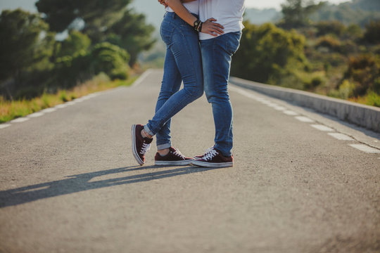 Crop side view of happy sensual couple in matching denim and sneakers embracing close while standing on countryside asphalt road at summer day