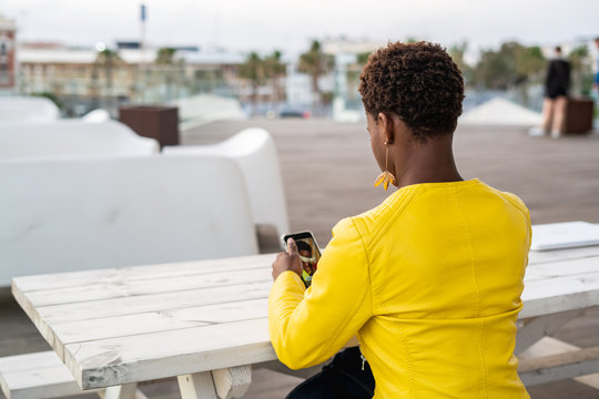 Back view of concentrated African American female messaging smartphone while relaxing at wooden table