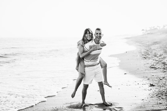 Full length cheerful adult male giving piggyback ride to smiling female while standing on wet sand near waving sea on resort and looking at camera