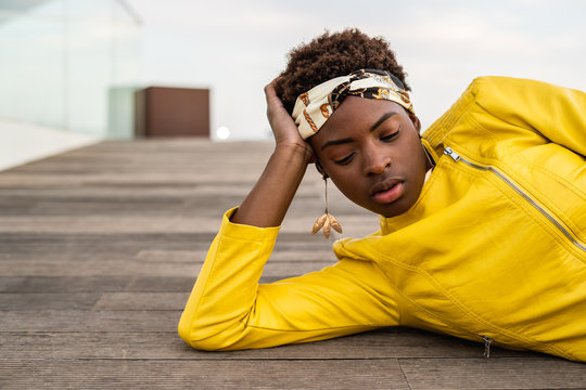 Stylish African American female in modern jacket relaxing lying down on wooden floor and looking down