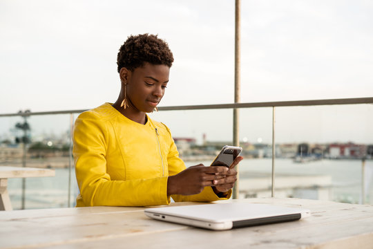 Concentrated African American female messaging smartphone while relaxing at wooden table