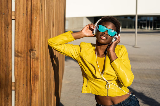 African American woman in stylish bright jacket and bright blue sunglasses using headphones standing near a modern building