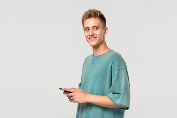 Smiling blond teenager in a green tee holding smartphone and looking at the camera isolated over light grey background.