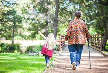 Rear view of senior woman walking with little girl in park. Grandmother walking with cane holding granddaughter with hand