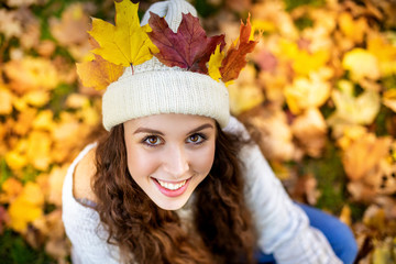 Young smiling woman in city park has colorful autumn leaves in hat and looking at camera