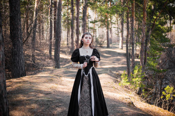 Fototapeta premium Young, beautiful girl in a black medieval dress with a steel rose in hand, walking through the forest.