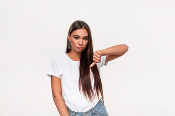Upset pretty african-american teenager in a white t-shirt and blue jeans holding thumbs down isolated over white background. Concept of dislike.