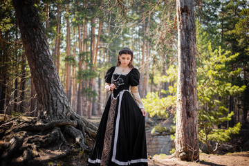 Young, beautiful girl in a black medieval dress with a steel rose in hand, walking through the forest.