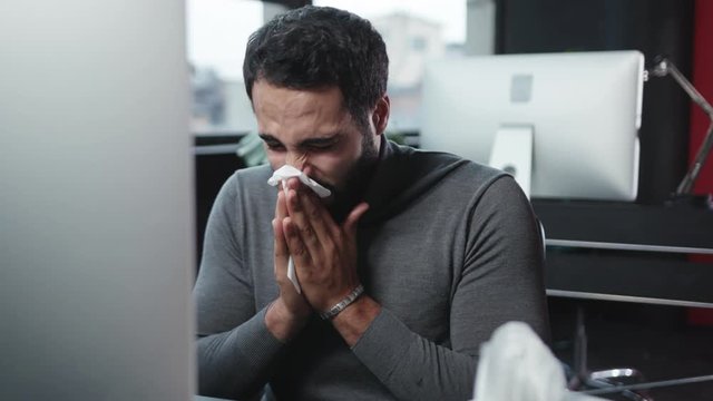 Sick bearded man in the office suffering fever or virus sneezing allergy and blowing his nose at desktop computer. Illness and tireness concept.