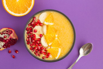 Smoothies bowl with orange, banana and pomegranate seeds on  the violet  background. Top view. Closeup.