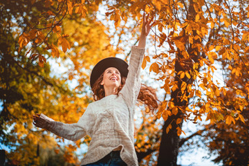 Smiling girl in hat jumping to colorful autumn leaves