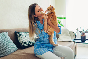 Playing with cat at home. Young woman sitting on couch teasing and hugging pet.