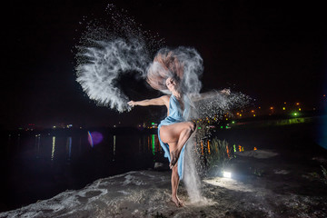 Red-haired woman in a blue dress with a deep neckline dancing in clouds of flour. A girl on the river bank jumping scattering white powder.