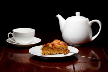 White tea cup and teapot with a slice of apple pie on a dark background from brown lacquered wooden boards. A logo can be conveniently placed on a cup and teapot.