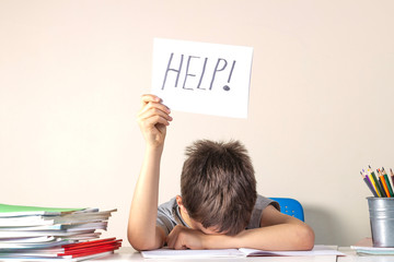 Sad tired frustrated boy sitting at the table with many books and holding paper with word Help....