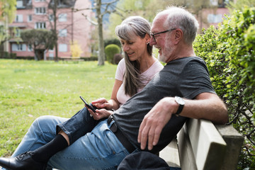 Happy senior couple using a phone sitting on a bench in the park
