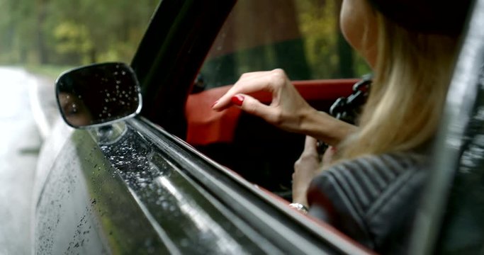 woman is sitting in luxury car parked on roadside in rainy day, smiling, view on face in rear mirror