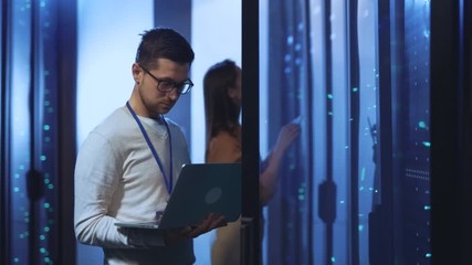 Data center IT specialist using laptop computer examining working rack network server cabinets. Two engineers inspecting server room at contemporary company. - Powered by Adobe