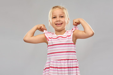 childhood and people concept - smiling little girl in striped dress showing her power over grey background