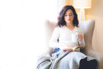 Young woman at home sitting on modern chair in front of window relaxing in her living room reading...