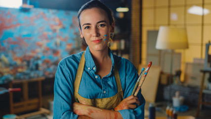 Young Female Artist Dirty with Paint, Wearing Apron, Crosses Arms while Holding Brushes, Looks at the Camera with a Smile. Authentic Creative Studio with Large Canvas. Head and Shoulders Portrait