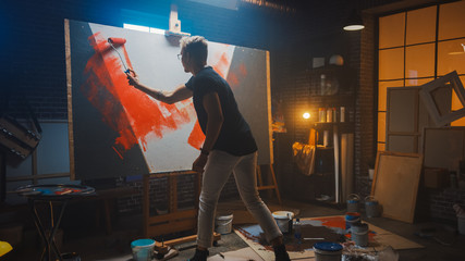 Talented Male Artist Working on an Abstract Painting, Uses Industrial Roller To Paint Daringly Emotional Modern Picture. Dark Creative Studio Large Canvas Stands on Easel Illuminated.