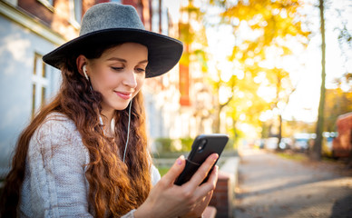 Beautiful stylish woman sitting on street at sunny day and using mobile phone with headphones