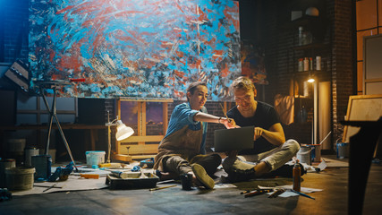 Couple of Artists Sitting in their Studio Use Laptop Computer, Working on a Project, Talking and Smiling. Painter and Her Partner Doing Research. Authentic Workshop with Oil Paintings and Tools