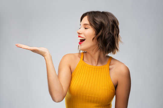 Emotions And People Concept - Happy Smiling Young Woman In Mustard Yellow Top Holding Something On Empty Hand Over Grey Background