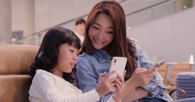 Little girl and her mother using smartphone at airport. Daughter talking to her mother with happy emotion.