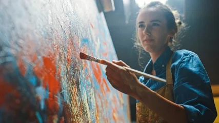 Tuinposter Female Artist Works on Abstract Oil Painting, Moving Paint Brush Energetically She Creates Modern Masterpiece. Dark Creative Studio where Large Canvas Stands on Easel Illuminated. Low Angle Close-up © Gorodenkoff