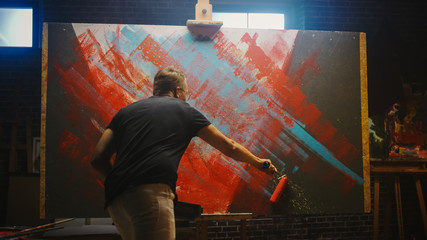 Talented Male Artist Working on a Abstract Painting, Uses Industrial Roller To Create Daringly Emotional Modern Picture. Dark Creative Studio Large Canvas Stands on Easel Illuminated