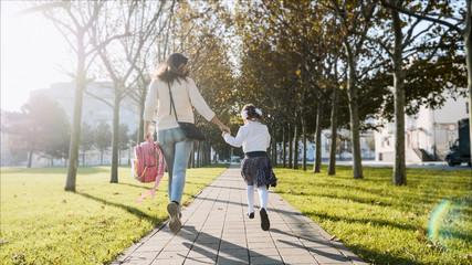 Young woman and little girl in school uniform are holding hands and running along the trees in the...