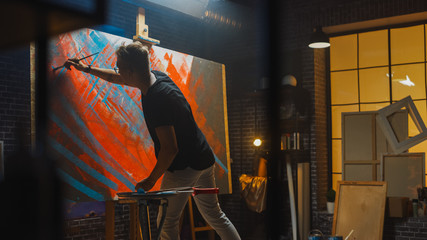 Talented male Artist Works on Abstract Oil Painting, with Broad Strokes of Paint Brush he Creates Modern Masterpiece. Dark and Messy Creative Studio where Large Canvas Stands on Easel Illuminated 