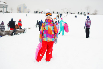 little girl in colored outerwear with a sledge child carriers in hands on the background of snow slope. Winter entertainment