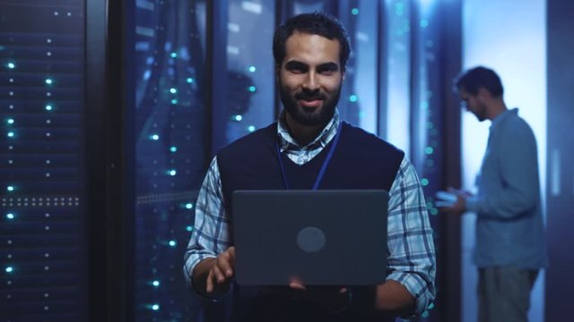 System administrator working with modern laptop in operative server rack room. Cheerful caucasian IT engineer in futuristic network data center. Development and success concept.
