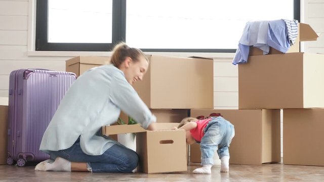 Little child looks in box. Background moving boxes and suit. Mother blonde woman wear blue shirt, jeans and white socks smile and look into box.