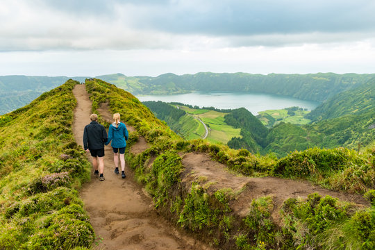 A Young Couple Holding Hands While Walking Towards The Grota Do Inferno Viewpoint At Sete Cidades On Sao Miguel Island, Azores. The Adults Are Walking Away From The Camera And Looking At The Lake View