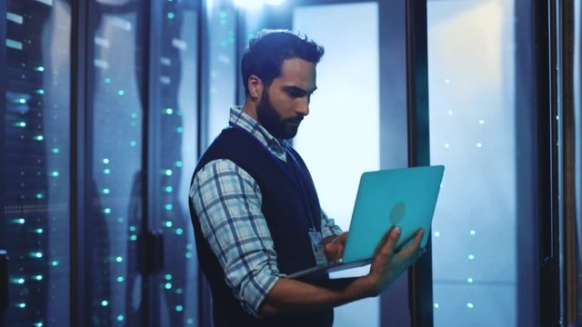 Server IT administrator working on laptop computer examining operation system of rack server cabinets in data center. Caucasian male technician in cyber security workspace.
