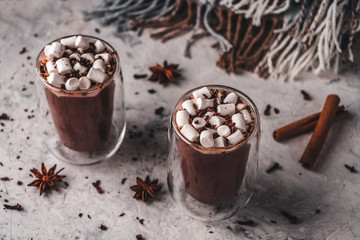 Two glasses of hot cocoa with marshmallows and chocolate chips on a background of scattered cloves, chocolate, star anise and cinnamon, with a blanket in the background