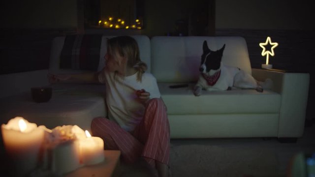 Funny girl watching TV program at evening , she hurries, jumps on sofa with a sandwich in hand, it is difficult to break away from the film. The dog is lying on the couch