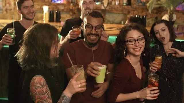 Medium shot of group of multiethnic young friends dancing with cocktails at bar or nightclub and smiling at camera