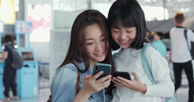 Mother talking to her daughter about travel trip at airport. Mother and girl using smartphone to searching information about travel trip together.