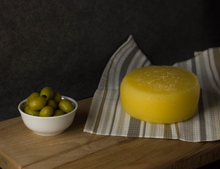 Cheese wheel on a napkin on a wooden Board, next to green olives in a plate.