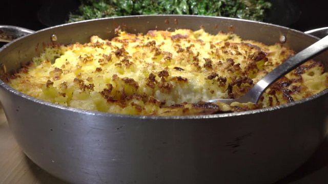 Normal speed followed by slow motion: close POV reverse dolly shot of a serving spoon taking the first portion of hot, baked, crispy topped, mashed potato and fish pie, from a pan.