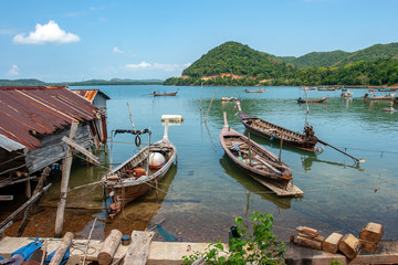 Fototapeta na wymiar Long fishing boats in Thailand on the water next to the fisherman's hut on stilts in the water. The old roof is very rusty. On the other side of the lake are overgrown green mountains.